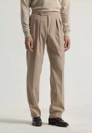 Pantalones plisados beige con cintura alta, cierre de botón y corte entallado. Combinados con un suéter beige claro y zapatos oscuros.