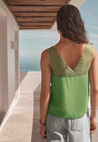 Green sleeveless top featuring a lace back detail, v-neck design, and smooth texture, paired with light blue denim jeans.