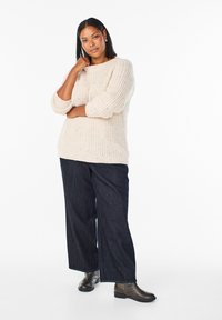 Pevný krémový pletený sveter s trojštvrťovými rukávmi, skombinovaný s voľnými tmavomodrými džínami a hnedými členkovými čižmami.
