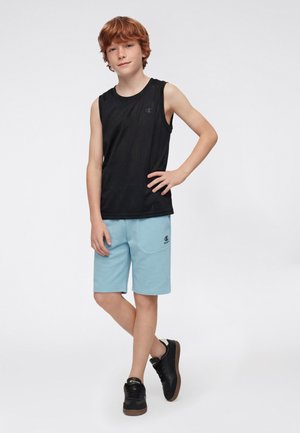 Chłopiec z rudymi włosami, ubrany w czarny bezrękawnik, jasnoniebieskie spodenki i czarne trampki, stoi z jedną ręką na biodrze na tle białego tła.