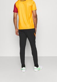 Kolorowy strój sportowy składający się z pomarańczowej i bordowej koszulki z krótkim rękawem oraz czarnych dopasowanych spodni, uzupełniony białymi i zielonymi butami piłkarskimi.