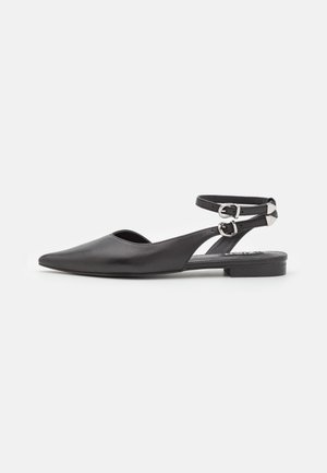 Chaussure plate noire à bout pointu avec doubles boucles argentées aux chevilles et côtés ouverts sur fond blanc.