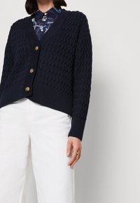 Marinblå stickad cardigan med en strukturerad mönster, V-ringning och tre bronsknappar. Kombinerad med vita byxor, som visar ribbade ärmslut och nederkant.