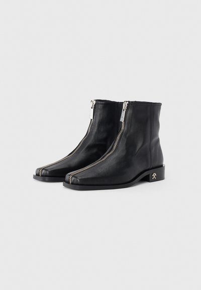 Botas de cuero negras hasta el tobillo con cremalleras frontales, punteras cuadradas, diseño minimalista y un tacón bajo en bloque que presenta un pequeño detalle decorativo.