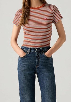 Kobieta ubrana w pomarańczowo-rdzawy i biały, pasiasty t-shirt z krótkim rękawem, wpuszczony do wysokich, ciemnoniebieskich dżinsów, z rękami w kieszeniach.