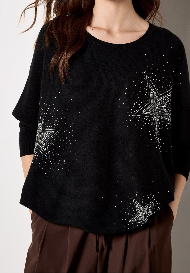 Haut en maille noir avec trois étoiles cloutées argentées, porté avec un pantalon marron, montrant le torse et les manches larges.
