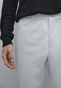 Hvide skræddersyede bukser med en glat overflade og subtile syninger, sammen med en mørk ribbet sweater. Nærbillede af hænder.