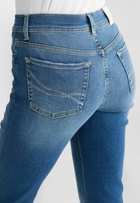Keskmise sinise pesu denim teksad, millel on vormitud disain, tagataskud, millel on rhinestone kaunistused, ja kõrge vöökoht.