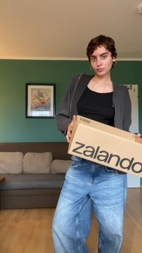 Smeđa kartonska kutija s natpisom "zalando" ispisanim crnom bojom. Osoba u sivoj jakni i crnom topu drži kutiju, uz svijetloplave traperice. Pozadina je zelena.