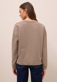 Kvinna vänd bort med ryggen iförd en taupe långärmad sweatshirt och mörkblå jeans mot en slät ljusbeige bakgrund.