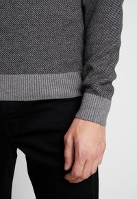 Grå pullovertröja med ett texturerat diagonalt mönster och ribbade muddar. Tyget verkar mjukt, med kontrasterande ljusare grått vid fållen.