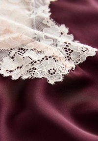 Burgunder-satin Stoff mit zarter weißer Spitzenverzierung, die blumige Muster und filigrane Details an den Kanten aufweist. Glatte Textur.