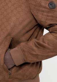 Καφέ φούτερ με σχέδιο και μαλακή υφή, με τσέπη με φερμουάρ, ριμπ μανσέτες και λεπτομέρεια με λογότυπο στο μανίκι.