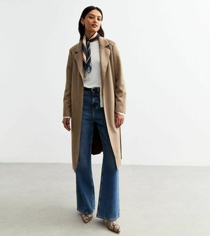 Manteau long beige sur une chemise blanche, associé à un jean large taille haute bleu et des talons à motifs. Une écharpe ajoute un détail à motifs.