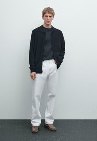 Zwarte cardigan over een donker shirt, witte losvallende broek en bruine loafers. Eenvoudig ontwerp, texturen die contrasteren tussen zacht en gestructureerd.