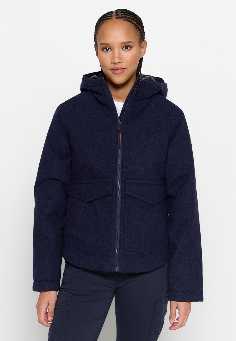 Giovane con i capelli intrecciati, che indossa una giacca a zip con cappuccio di colore blu navy e pantaloni cargo scuri, in piedi contro uno sfondo bianco.
