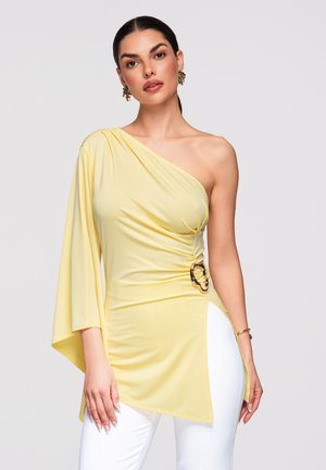 Tummatukkainen nainen, jolla keltainen epäsymmetrinen yhden olkapään toppi ryppykuvioinnilla ja valkoiset housut, poseeraa yksinkertaisen taustan edessä.