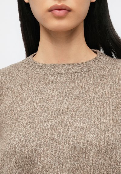 Femme aux cheveux longs et foncés portant un pull beige en maille à col rond, cadrée pour montrer le cou et le haut de la poitrine.