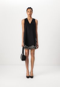 Black sleeveless dress featuring a V-neck, scalloped white trim at the hem, and a relaxed fit. Worn with black high heels and a small handbag.