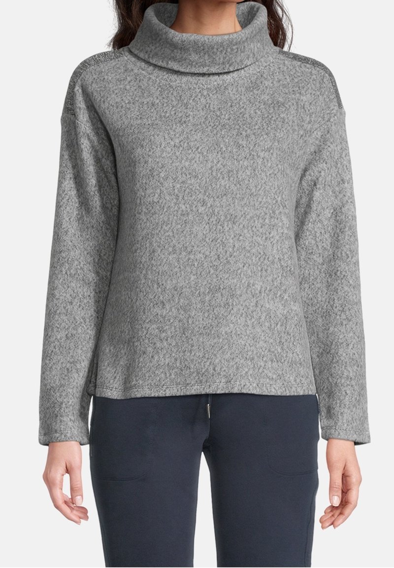 Femme portant un pull polaire gris à manches longues avec un col roulé ample et un pantalon foncé, debout devant un fond uni.