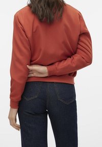 Blouson aviateur en satin rouge avec poignets côtelés, porté sur un jean denim bleu foncé, présentant une coupe décontractée et une texture lisse.