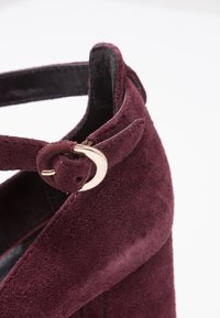 Gros plan sur une chaussure compensée en daim bordeaux avec une lanière pour la cheville maintenue par une petite boucle dorée.