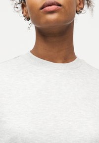Gros plan sur le bas du visage et le cou d'une personne aux lèvres naturelles, portant de petites boucles d'oreilles argentées en forme d'anneau et un t-shirt gris clair à col rond.