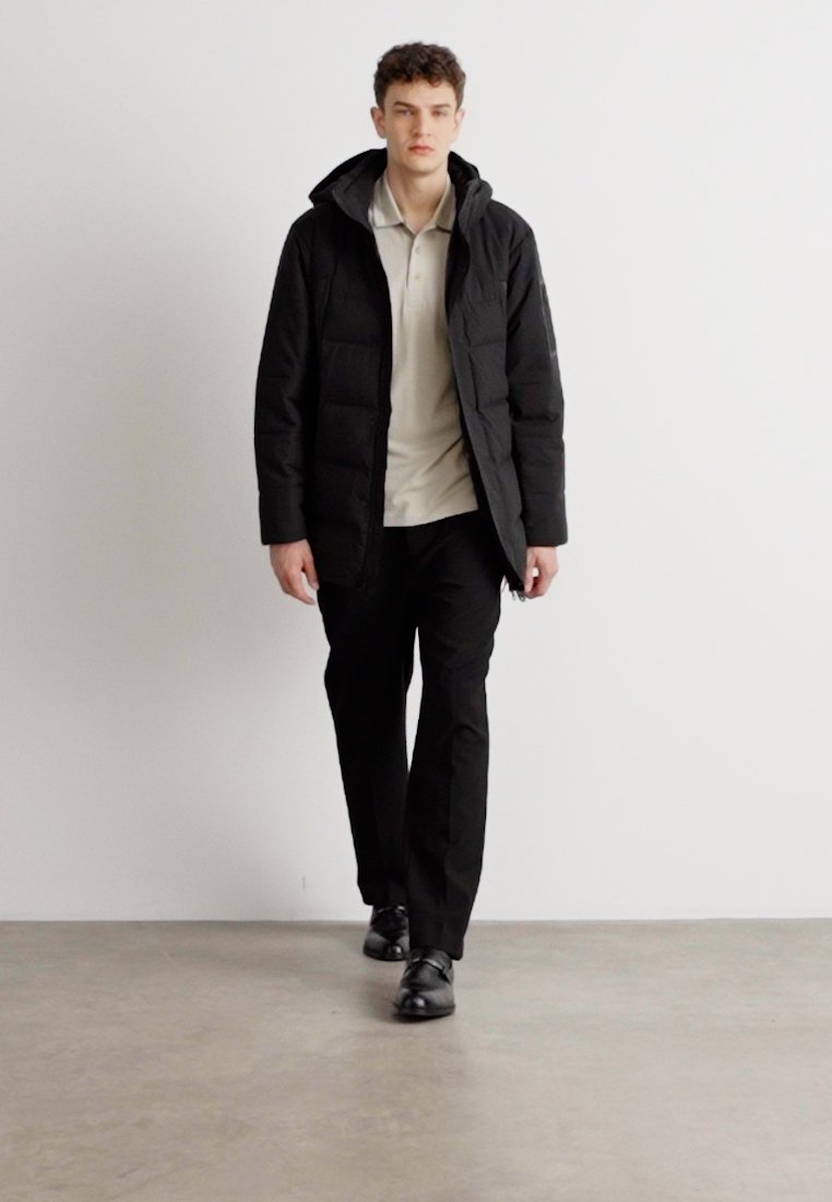 Parka noire avec une capuche, présentant un design matelassé. Portée sur une chemise polo de couleur claire et un pantalon noir, complétée par des chaussures noires.
