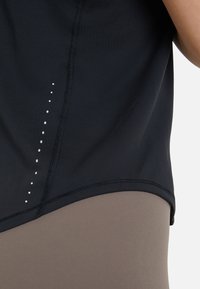 Črna športna majica z teksturiranim zaključkom, z ravnim robom in vertikalnimi belimi pikami. V kombinaciji z bež leginsami.