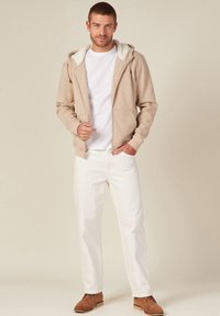 Felpa leggera beige con cerniera e cappuccio foderato in grigio, indossata sopra una t-shirt bianca, abbinata a jeans bianchi e scarpe in suede marroni.