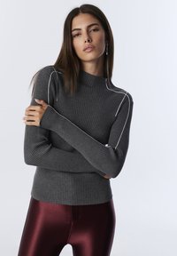 Young woman with long hair wearing a dark gray ribbed sweater with white stitching and shiny burgundy leggings against a plain background.