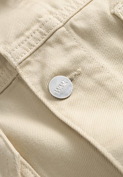 Sidabrinis mygtukas su graviravimu „W.W.“, pritvirtintas prie smėlio spalvos džinsinio audinio su matomomis siūlėmis ir siūlėmis.