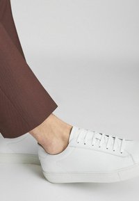 Fehér bőr sportcipő, hat fűzőlyukkal és arany logós díszítéssel, barna slim-fit nadrággal párosítva, amely fedi a boka területét.