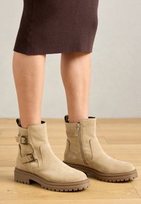 Bottines en daim beige avec deux boucles latérales et une fermeture éclair. Semelles en caoutchouc brun épais avec une texture nervurée pour une meilleure adhérence.