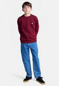 Vínová mikina s rebrovanými manžetami, kombinovaná s voľnými modrými rifľami. Čierne tenisky s gumovou podrážkou dopĺňajú outfit.