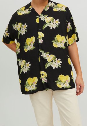 Mand iført kortærmet sort skjorte med knapper med gul citron og hvid blomstermønster, kombineret med lyse beige bukser.