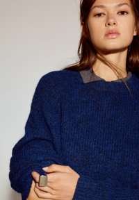 Donkerblauwe trui met een textuur gebreid patroon, losse pasvorm en geribbelde boorden, gedragen over een grijs overhemd met kraag. Zilveren ring aan de hand.