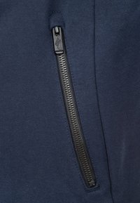 Marinblå ziphoodie med texturerad tyg, med svart dragkedjestängning och en liten logotyp på dragfliken.