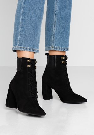 Stopy w czarnych, zamszowych botkach na obcasie z sznurowadłami i złotymi przelotkami, w połączeniu z jasnoniebieskimi, skróconymi jeansami na jednolitym tle.