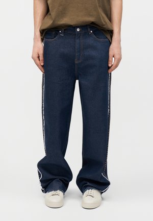 Sötétkék, széles szárú farmernadrágot viselő személy, fehér oldalszegélyű varrással és fehér tornacipőben, egyszínű háttér előtt állva.