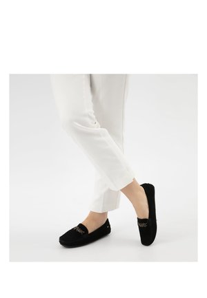 Persona che indossa pantaloni bianchi e mocassini neri senza lacci con dettaglio a catena decorativa, in piedi con un piede leggermente sollevato.