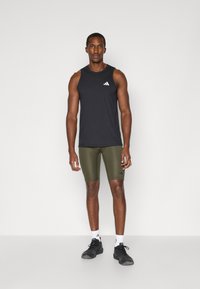 Top deportivo sin mangas negro con logo blanco, combinado con pantalones cortos ajustados en verde oliva. El modelo lleva zapatillas negras y calcetines blancos.