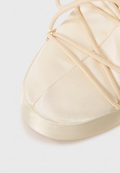 Primo piano di una ballerina in satin beige con fasce elastiche incrociate e punta arrotondata su uno sfondo bianco.