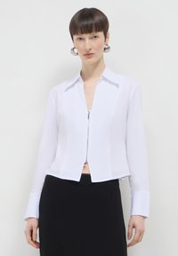 Maglietta bianca con zip, maniche lunghe, colletto e design cropped. Il tessuto appare liscio; presenta un aspetto pulito e sartoriale.