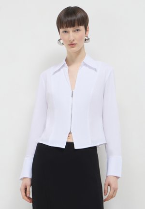 Chemise blanche zippée avec manches longues, col et un design cropped. Le tissu semble lisse et présente une apparence propre et ajustée.