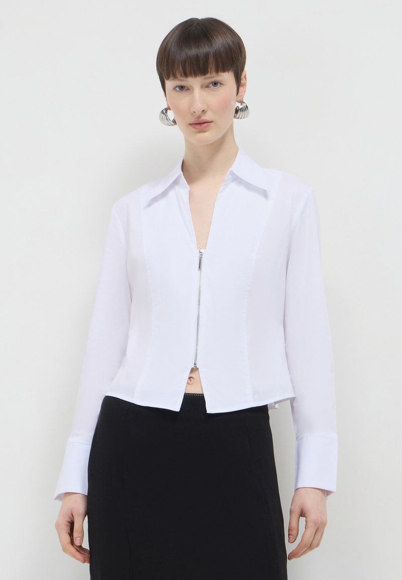 Maglietta bianca con zip, maniche lunghe, colletto e design cropped. Il tessuto appare liscio; presenta un aspetto pulito e sartoriale.