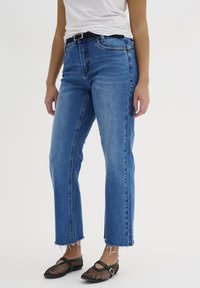 Jeans de mezclilla azul con un diseño de corte recto, cintura alta y dobladillo deshilachado. Llevados con una blusa blanca lisa y planos de malla negra.