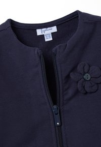 Marineblauer Baumwoll-Cardigan mit Reißverschluss, versehen mit einem Stoffblütenakzent und einem Etikett, das die Größe 12-18 Monate angibt.