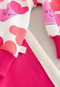 Hanorac colorat cu modele de inimioare roz și roșii pe material alb, cu manșete striate și o etichetă mică „Little Bird”. Strat inferior roz.