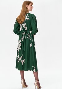 Zielona sukienka w kwiatowy wzór z długimi rękawami, zawiązana w talii. Gładka tkanina z długością do połowy łydki, w zestawieniu z jasnymi złotymi szpilkami.